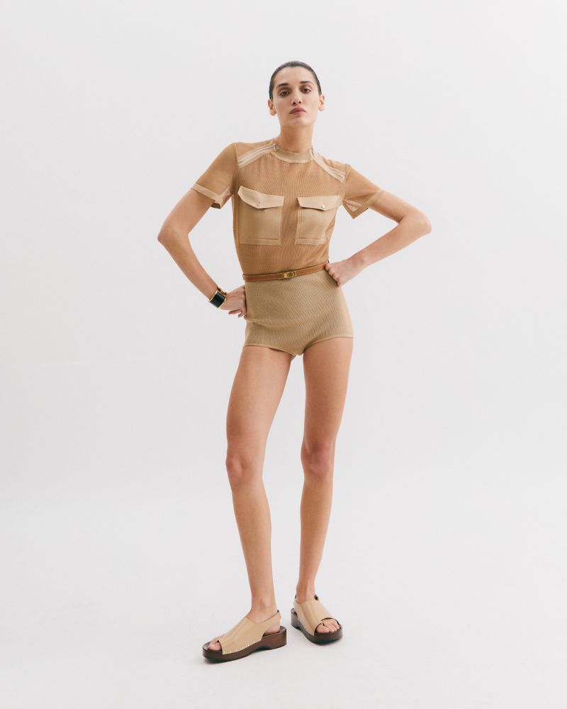 A woman poses in a tan, sheer short-sleeved top, tan shorts, and chunky sandals against a white background in a fashion forward pose.