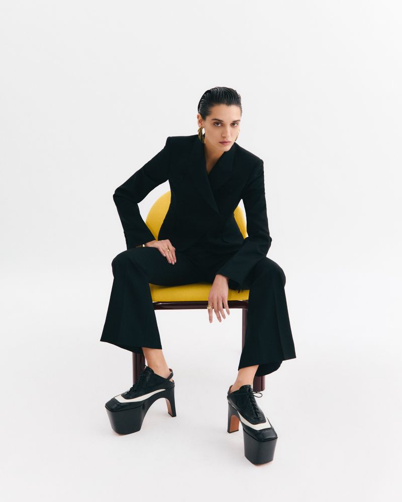 Stylish woman in a black pantsuit sitting on a yellow chair against a white backdrop, showcasing platform heeled shoes and gold earrings.