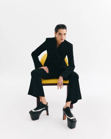 Stylish woman in a black pantsuit sitting on a yellow chair against a white backdrop, showcasing platform heeled shoes and gold earrings.