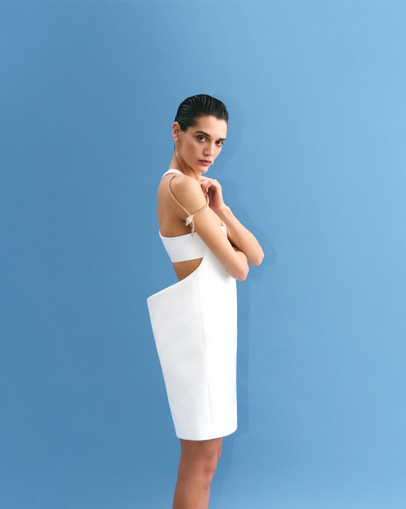 Woman in white dress poses against a light-blue backdrop in a fashion shot with studio lighting, cropped to a medium view.