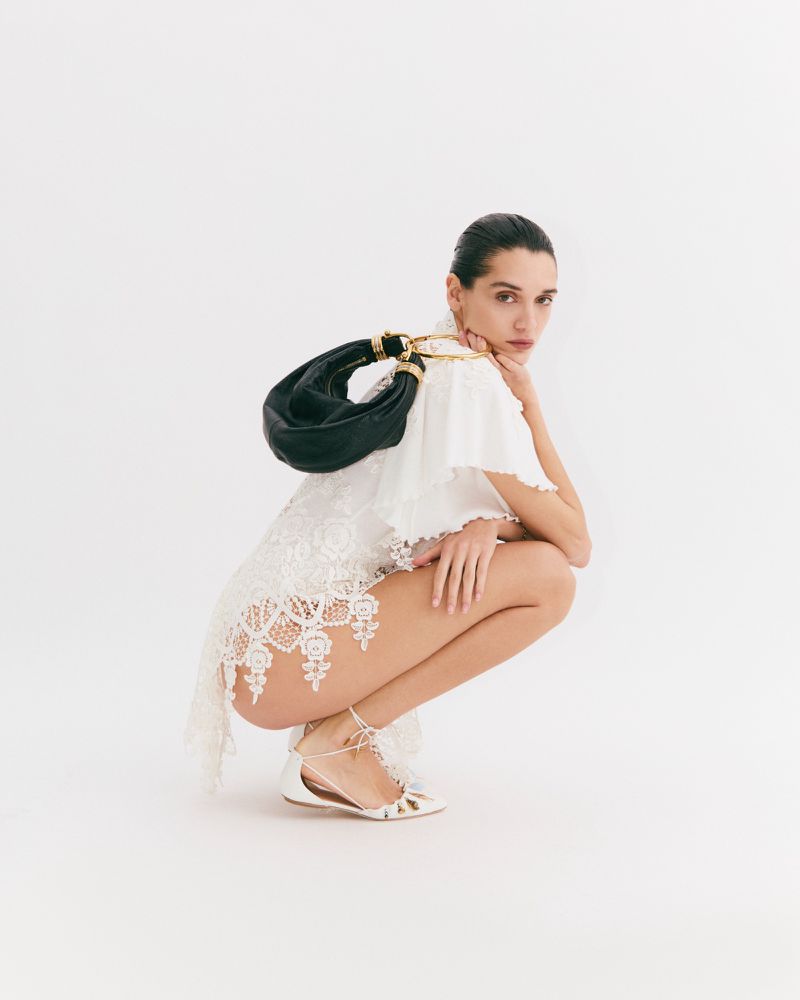 A woman wearing a white lace dress and white sandals, squatting, with a black leather handbag against a white background.