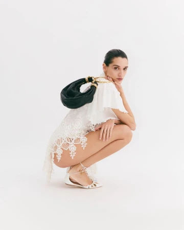 A woman wearing a white lace dress and white sandals, squatting, with a black leather handbag against a white background.