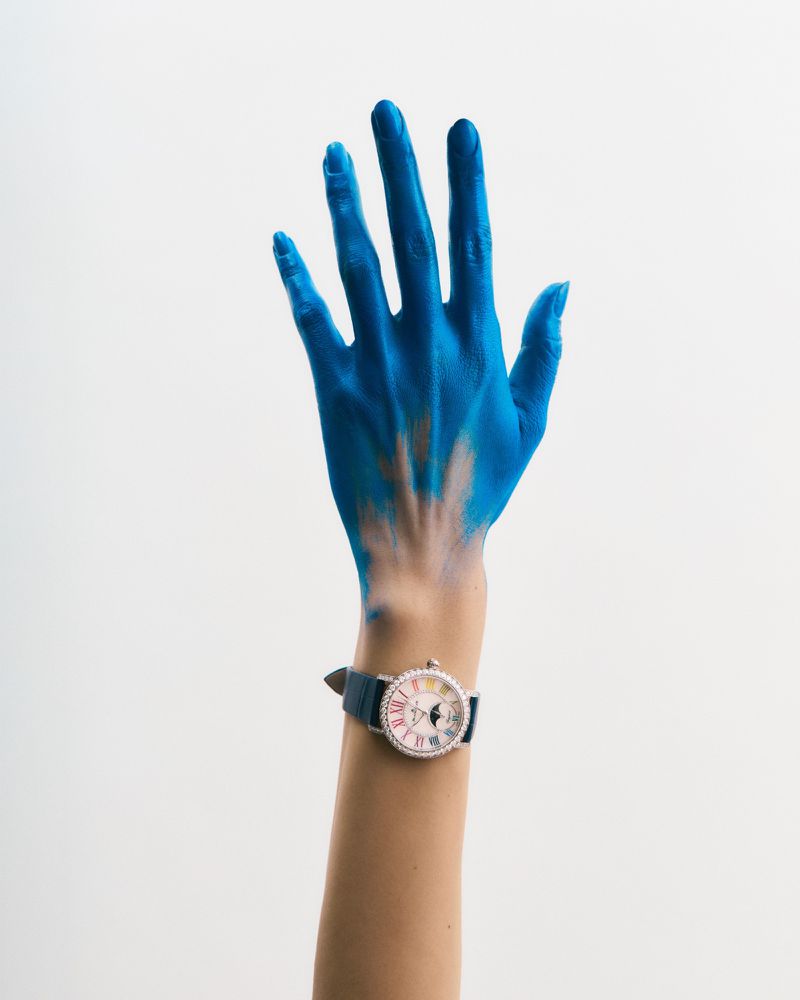 Arm raised with blue paint on hand and wrist watch against white backdrop. Clean studio-style shot, showing vibrant color contrast.