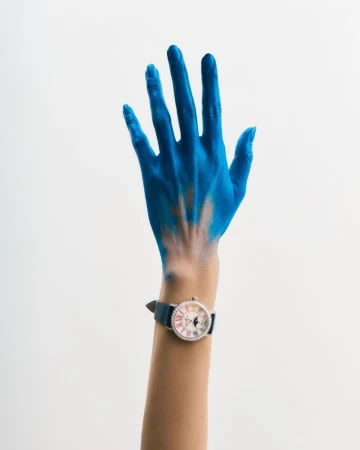 Arm raised with blue paint on hand and wrist watch against white backdrop. Clean studio-style shot, showing vibrant color contrast.