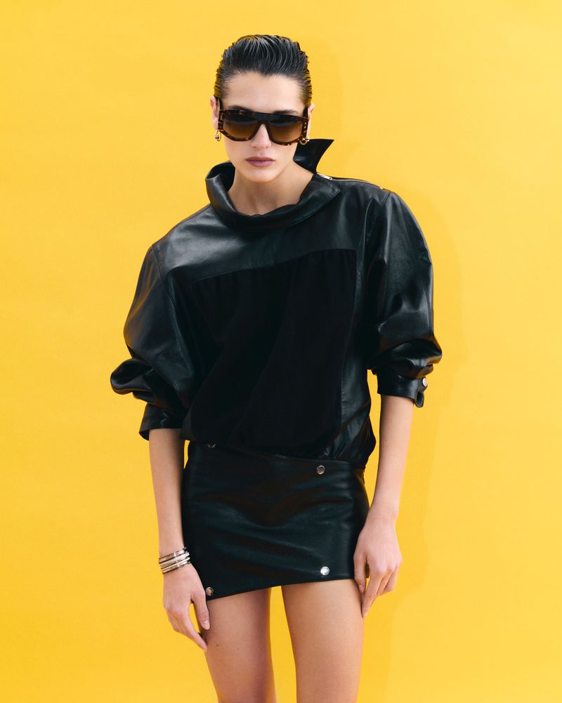 Studio shot of a woman with sunglasses posing in a black leather top and skirt against a yellow background, showcasing fashion apparel.
