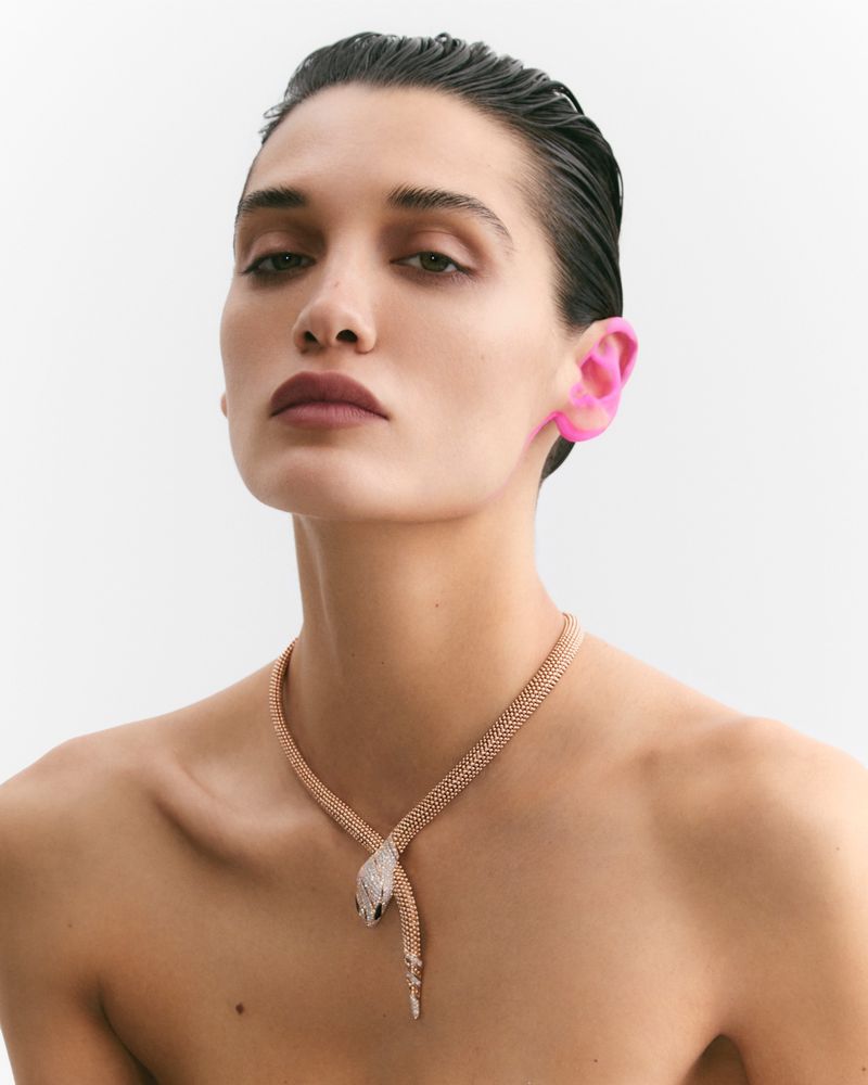 Close-up of a model with pink ear prosthetics and a snake necklace. The model is in front of a bright white background.