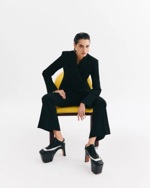 Woman wearing a black suit and chunky platform shoes seated on a yellow chair against a white background.