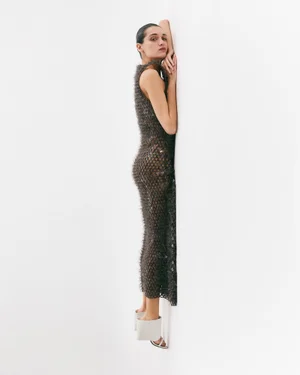 Model in dark textured sleeveless dress leaning against a plain white wall wearing white mules