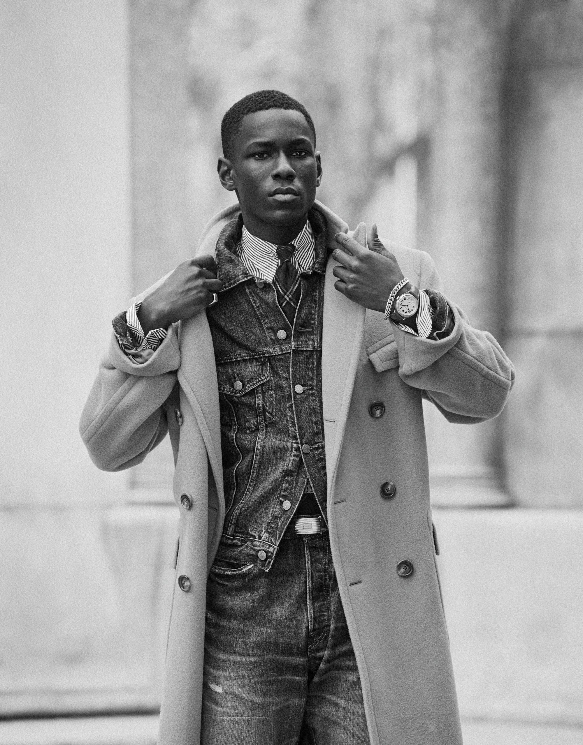 Black man wearing a layered coat and denim jacket against an urban architectural backdrop in New York