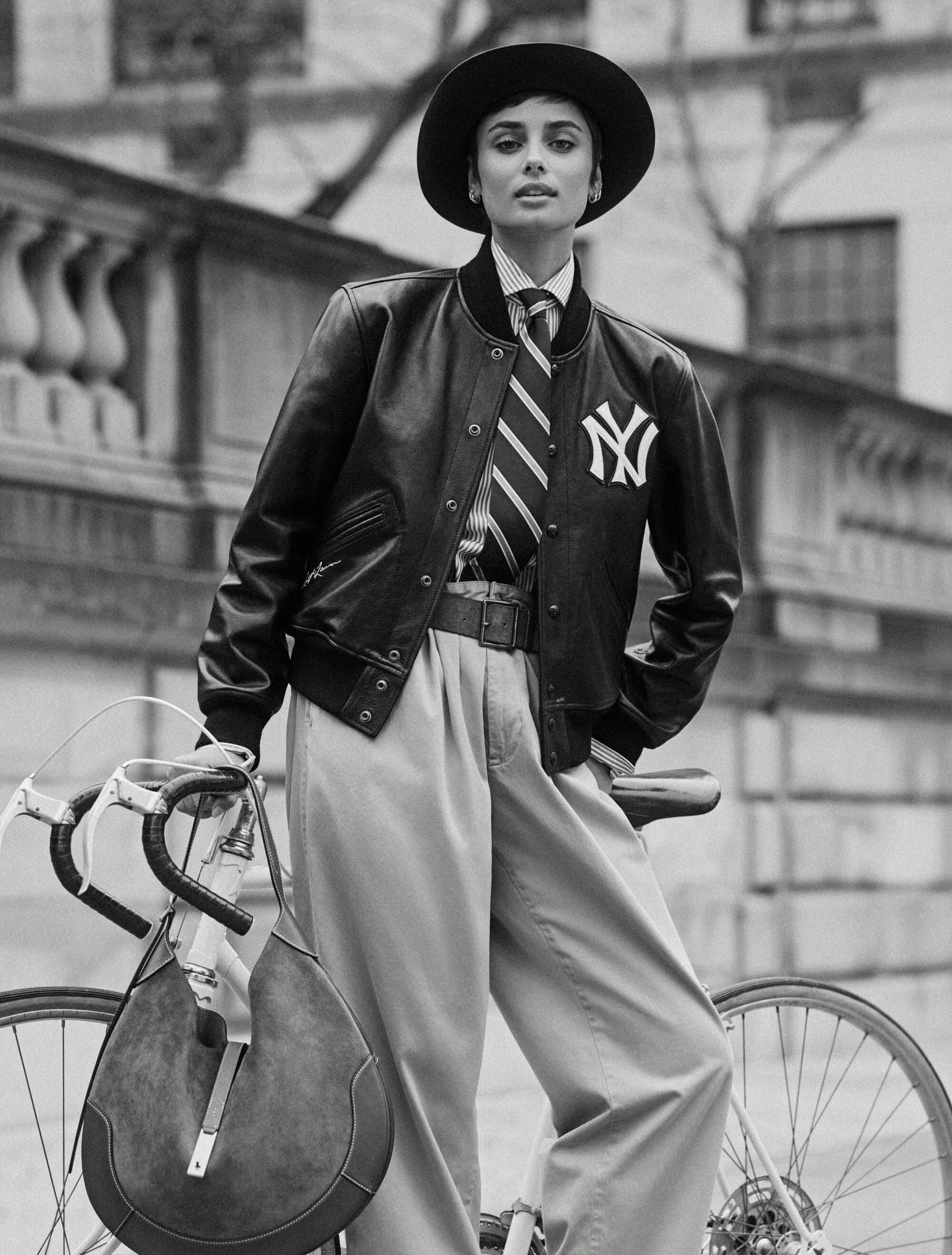 Model wearing a leather jacket, striped tie, wide trousers, and a wide-brim hat, holding a bicycle and handbag.