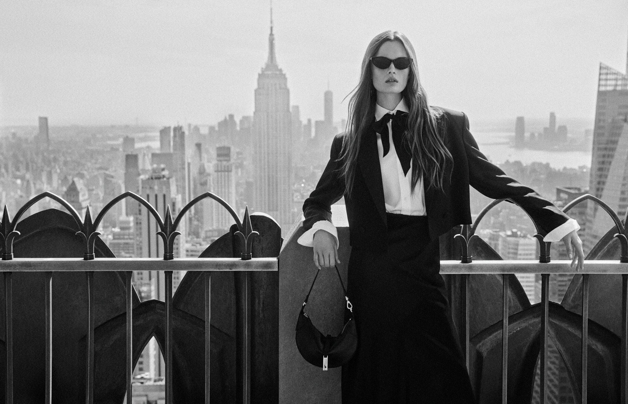 A woman wearing dark sunglasses, black blazer, white shirt with bow tie, and holding a handbag on a New York rooftop.
