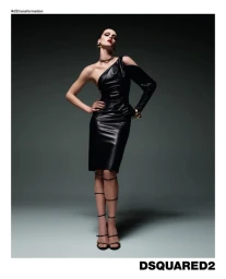 Model Abby Champion wearing a black leather one-shoulder dress and gloves against a dark gradient studio backdrop.