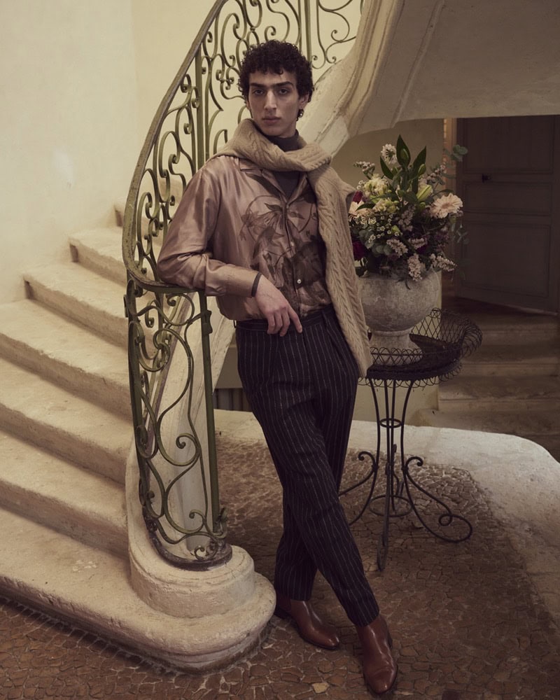 A man wearing a patterned shirt, dark trousers, and a scarf leaning against an ornate iron staircase railing inside a hall.