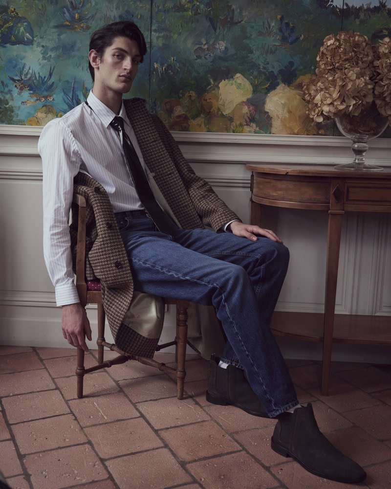 Man seated on a wooden chair wearing a white shirt, tie, and draped patterned jacket for Baldessarini campaign.