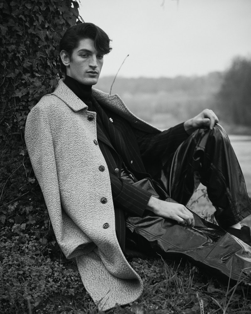 Man wearing a textured coat and dark trousers seated outdoors against an ivy-covered tree trunk in a black and white portrait.