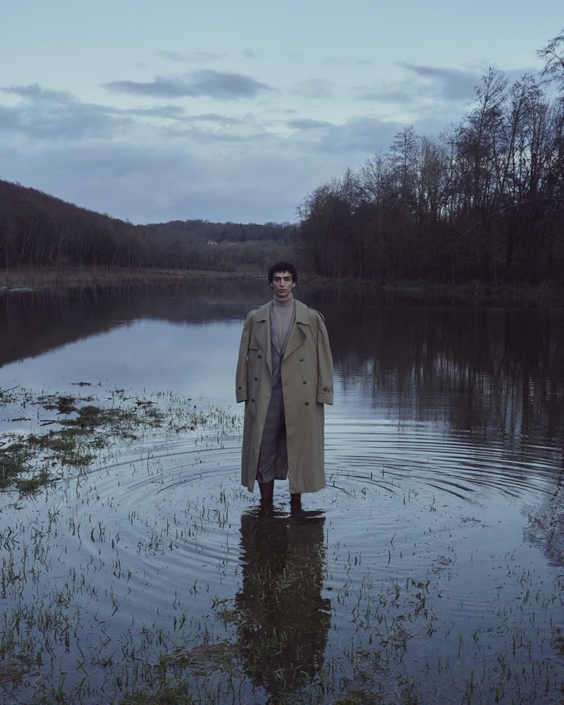 Man wearing a long beige trench coat standing ankle-deep in calm water with dark trees under an overcast sky.