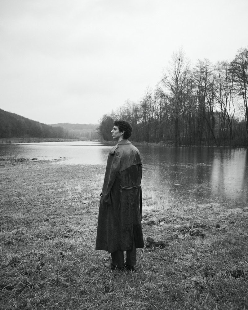 Man wearing a long dark coat standing outdoors in a wet landscape by a lake, photographed in monochrome.