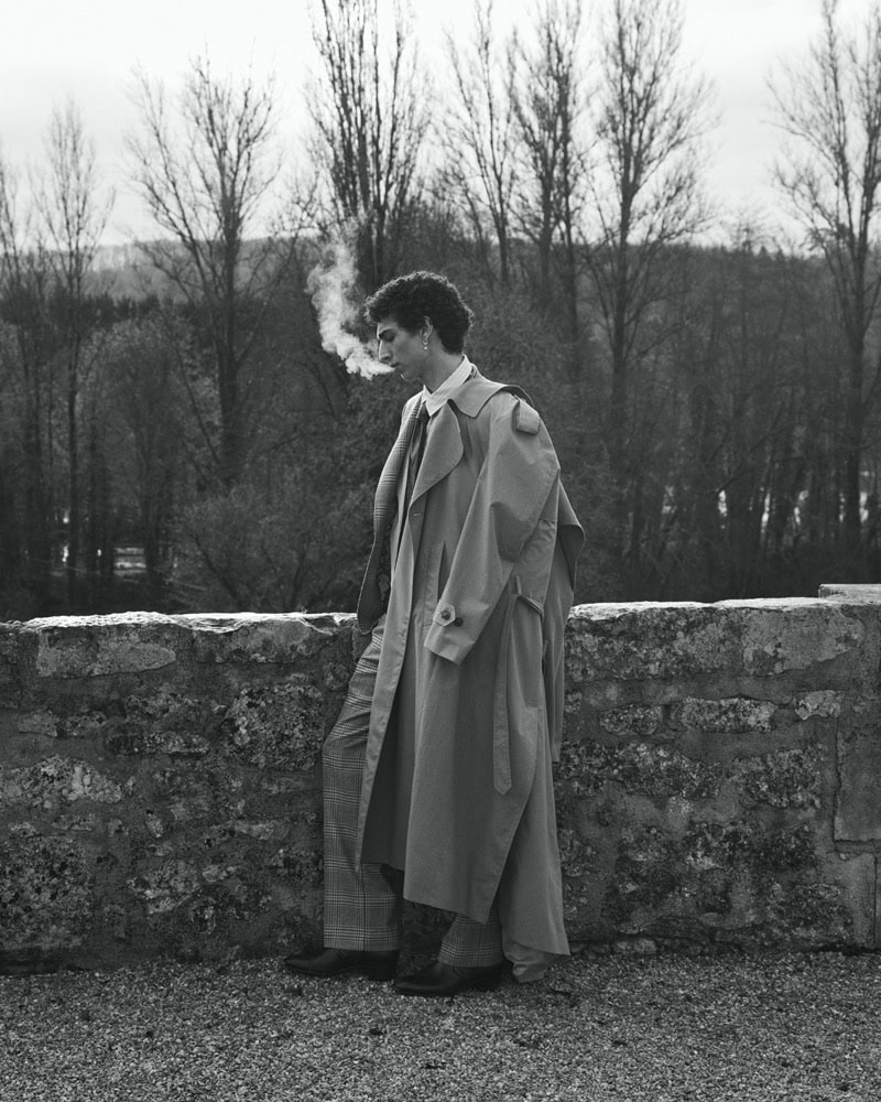 Man wearing a long trench coat leaning against a stone wall outdoors with bare trees in the background.