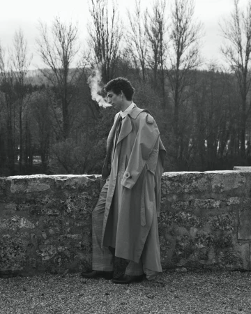 Man wearing a long trench coat leaning against a stone wall outdoors with bare trees in the background.