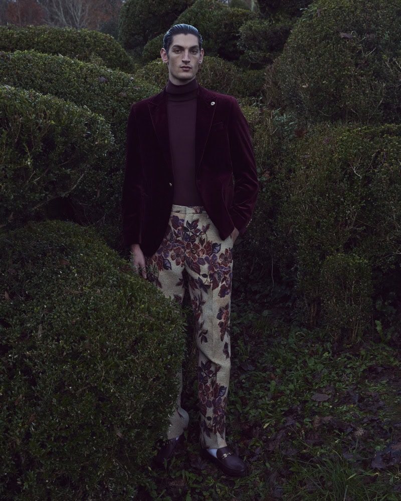 Model standing among sculpted topiary hedges wearing a burgundy velvet blazer and floral trousers