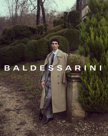 Man wearing a long beige trench coat over a grey suit standing outdoors near trimmed hedges and topiary trees.