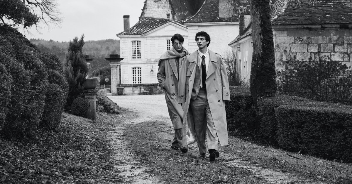 Two male models in trench coats walking along a gravel path in front of a classical French estate in black and white.