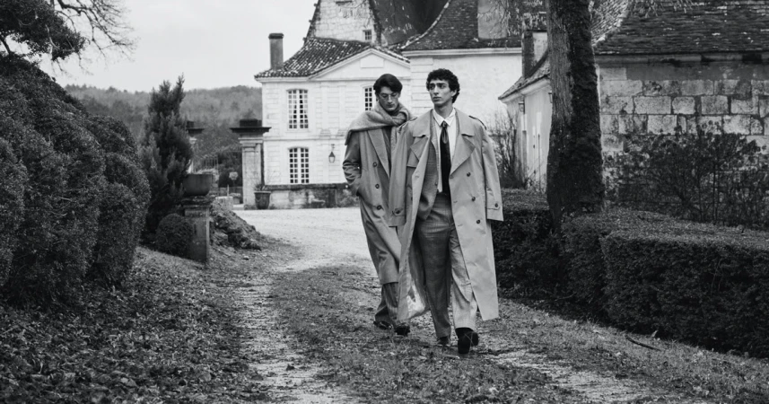 Two male models in trench coats walking along a gravel path in front of a classical French estate in black and white.