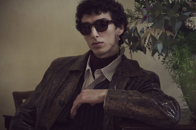 Man wearing dark sunglasses and brown jacket seated near a floral arrangement
