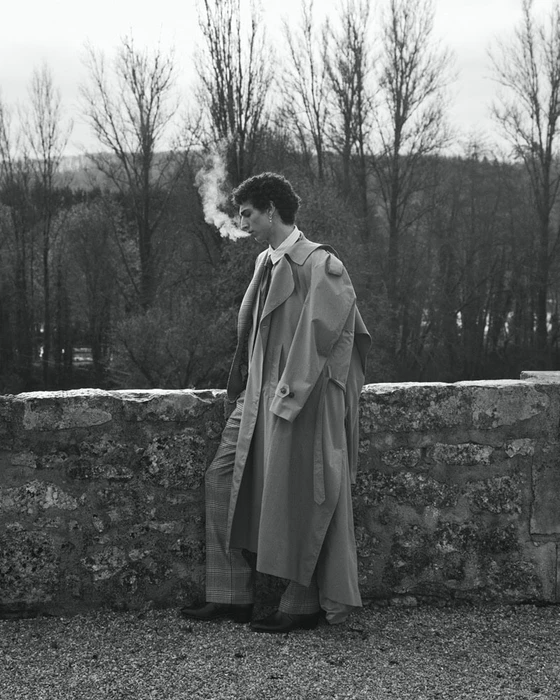 Man wearing a long trench coat leaning against a stone wall outdoors with bare trees in the background.