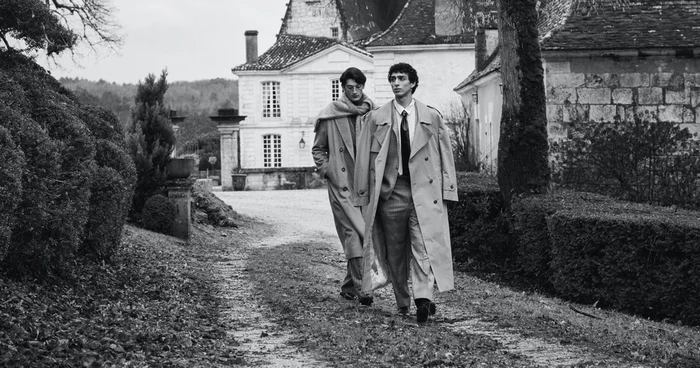 Two male models in trench coats walking along a gravel path in front of a classical French estate in black and white.