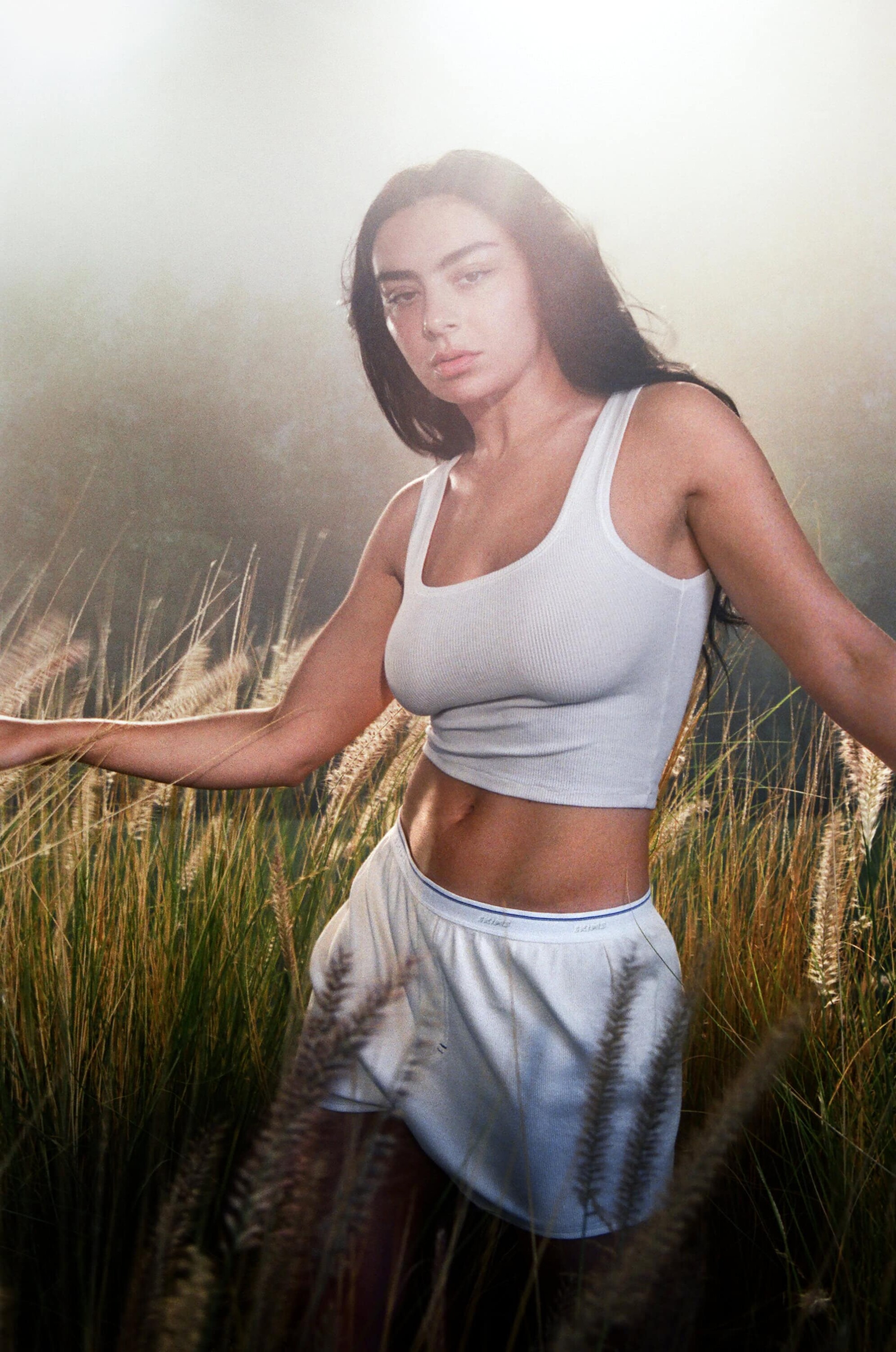 Woman wearing a white tank top and white shorts posing in tall grass under bright sunlight.