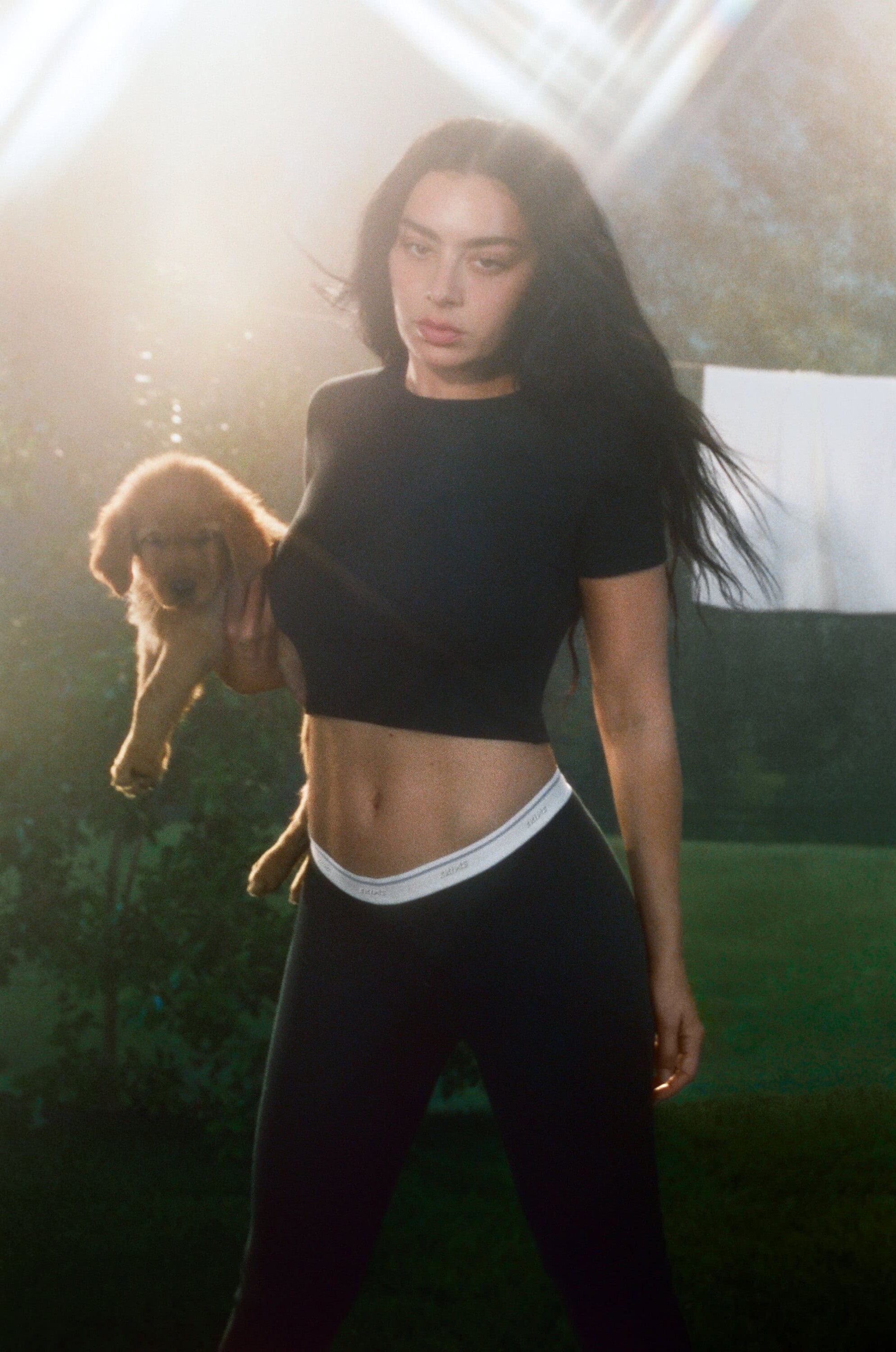 Woman with long dark hair wearing a black crop top and black leggings holding a small puppy outdoors.
