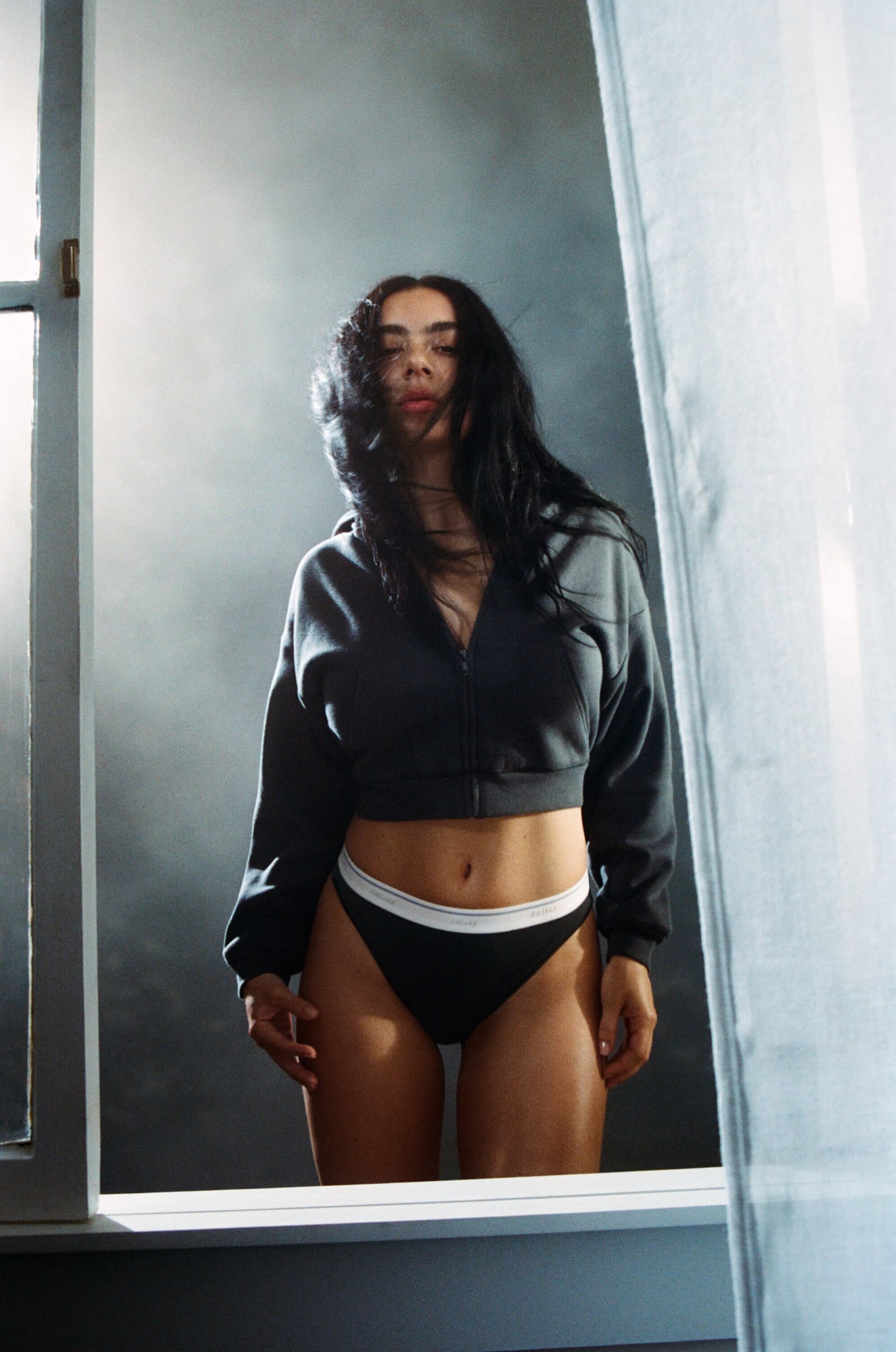 Woman with long dark hair wearing a cropped grey zip-up hoodie and dark underwear against a cloudy grey background.