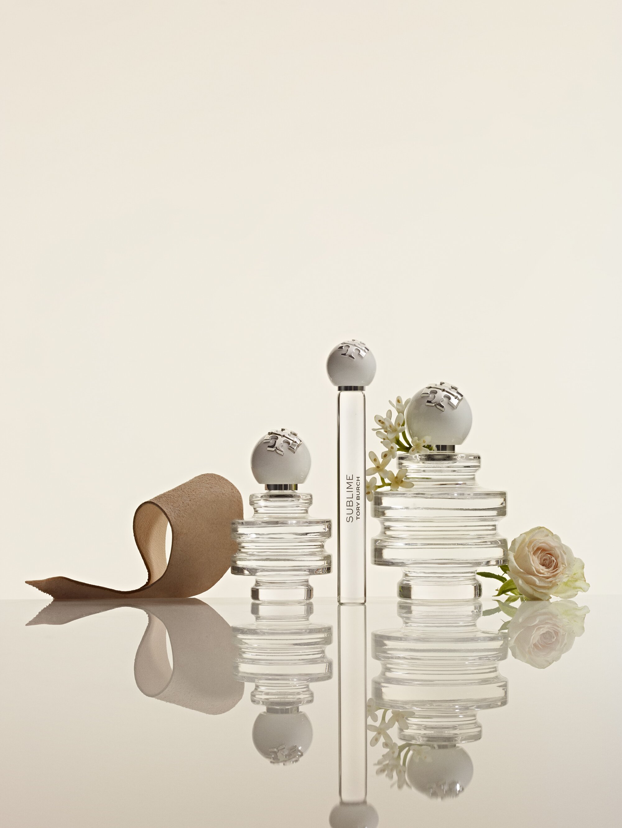 Tory Burch perfume bottles with spherical stoppers displayed on a reflective white surface alongside a beige ribbon and white flower.