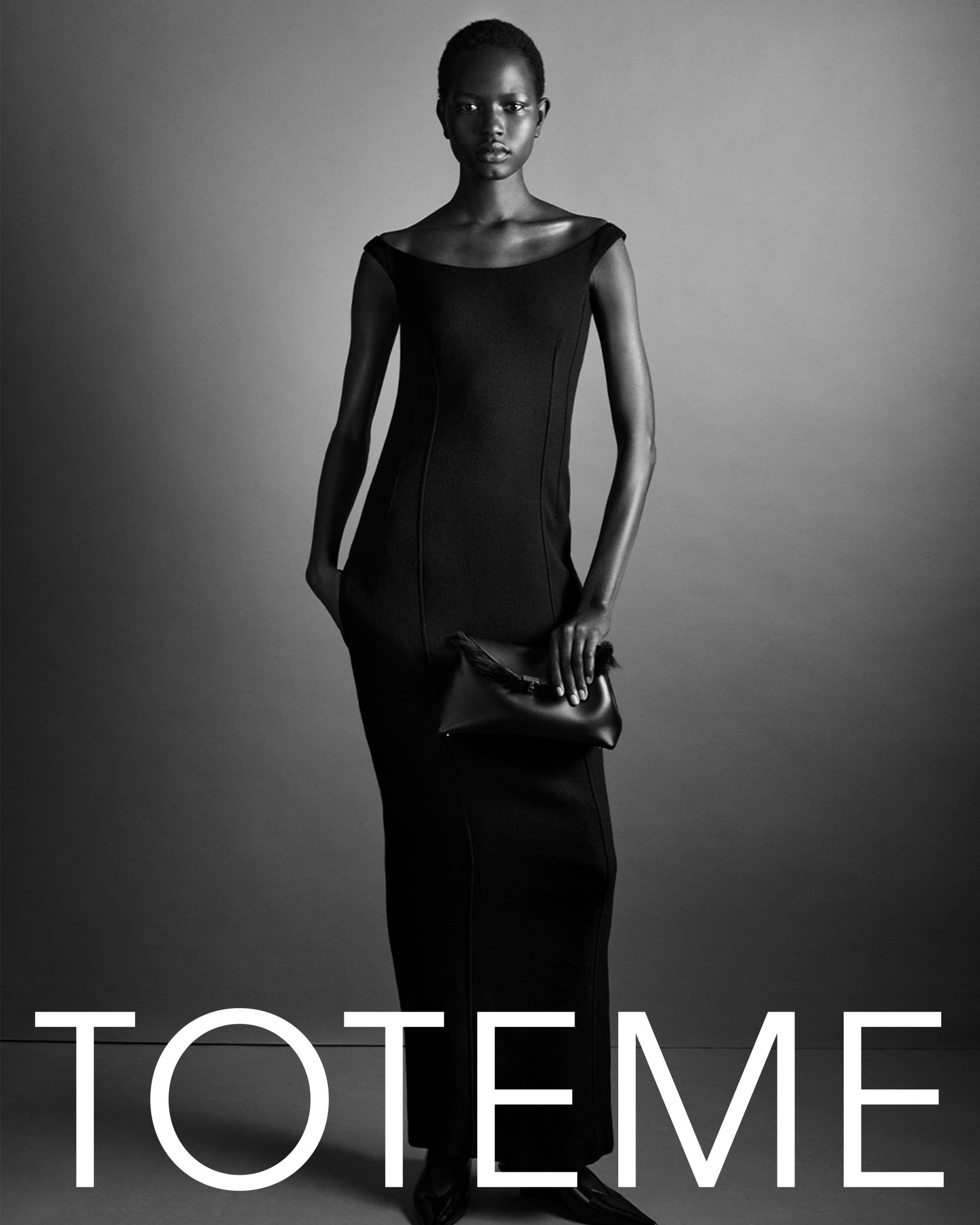 Black and white studio portrait of a model in a long dark dress holding a black clutch for Toteme