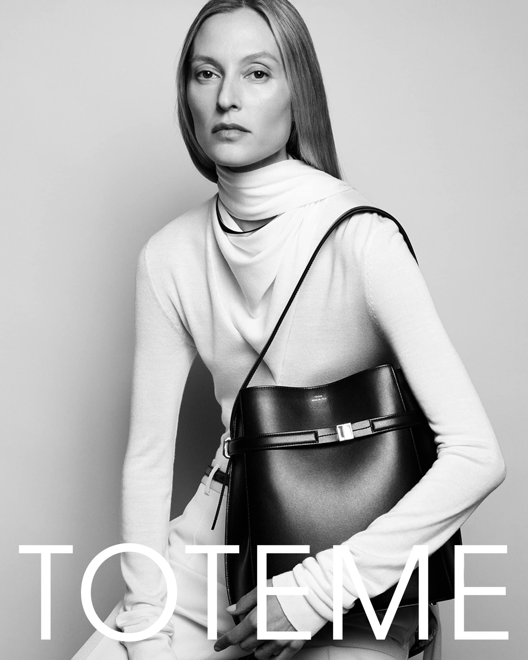 Model in white turtleneck top holding a black leather shoulder bag against a grey seamless studio background for Toteme fall 2024 campaign