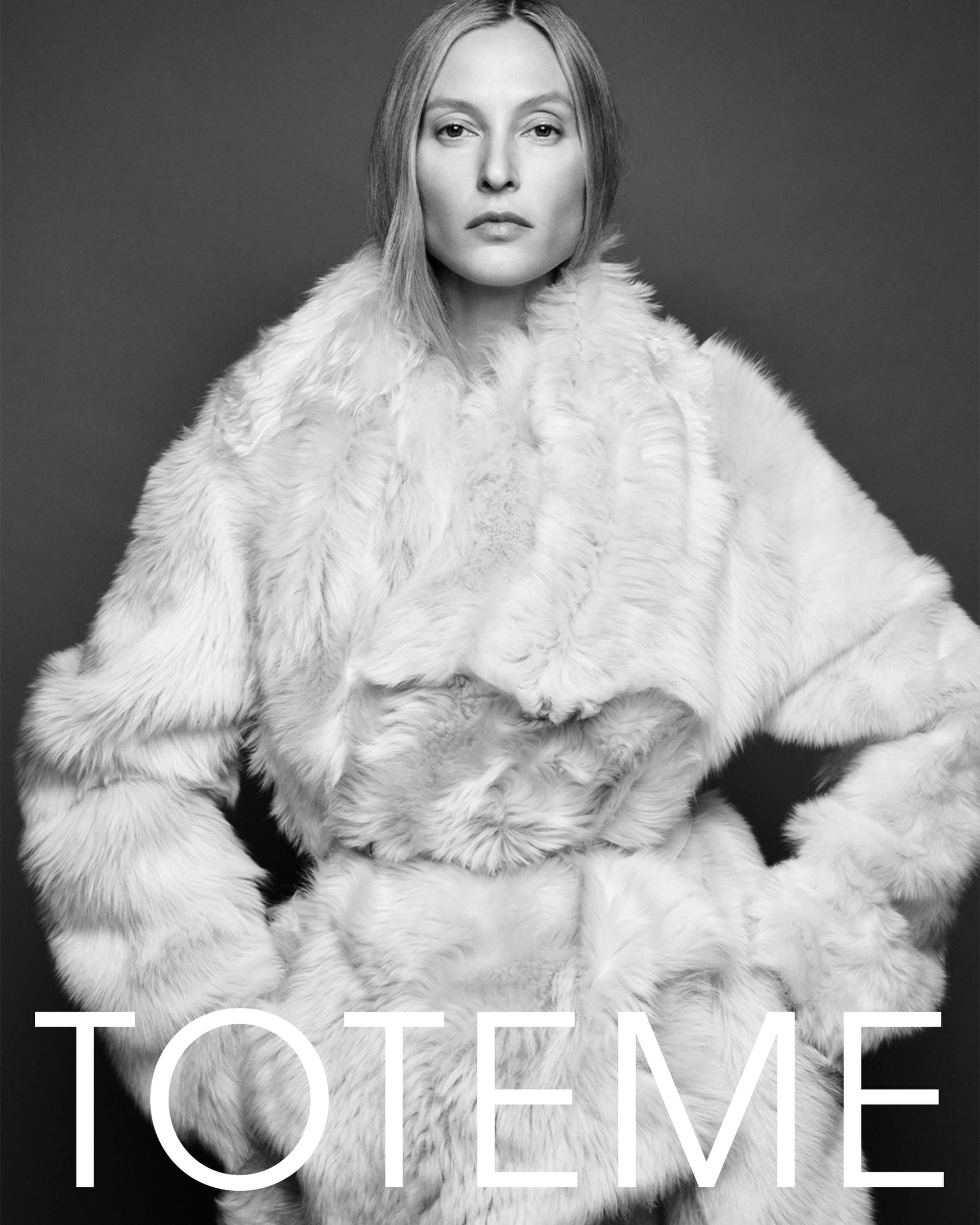 Model wearing a textured white fur coat against a solid dark grey studio background for Totême Fall 2024 ad campaign.