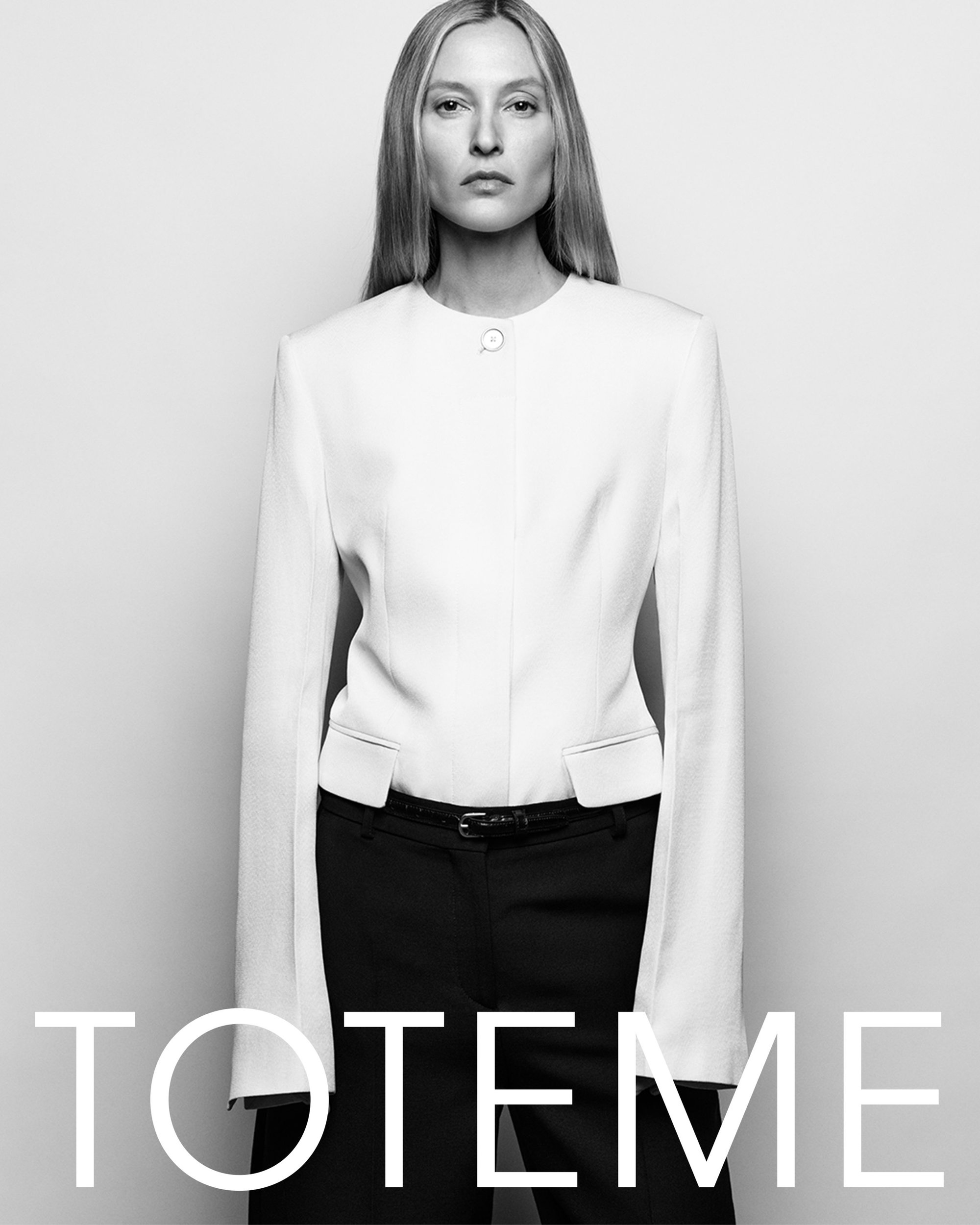 Model in white collarless jacket and dark trousers against a plain studio background with Toteme branding.