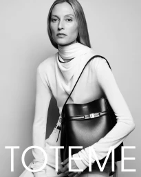 Model in white turtleneck top holding a black leather shoulder bag against a grey seamless studio background for Toteme fall 2024 campaign