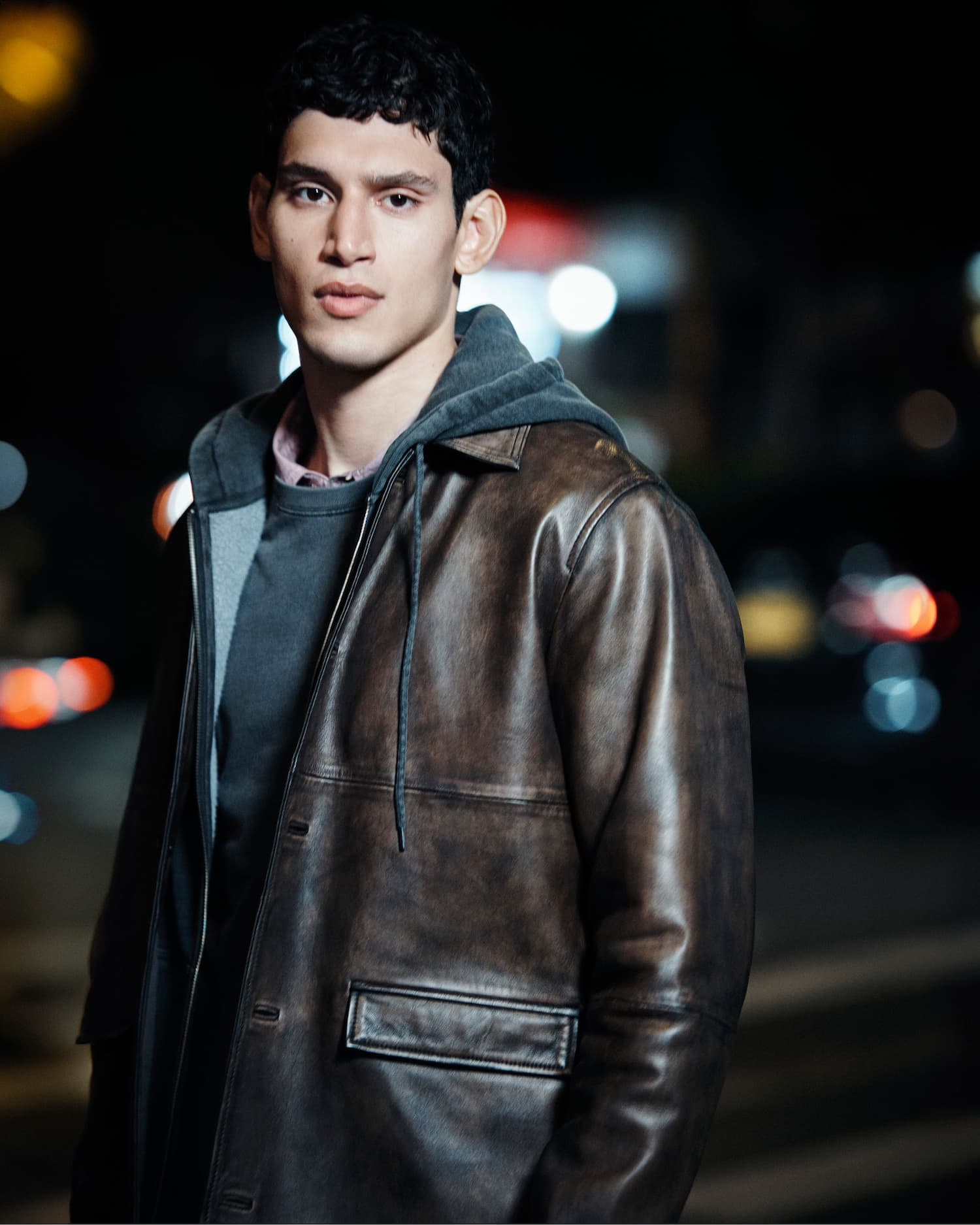 Man wearing a distressed brown leather jacket and grey hoodie against a dark nighttime street background