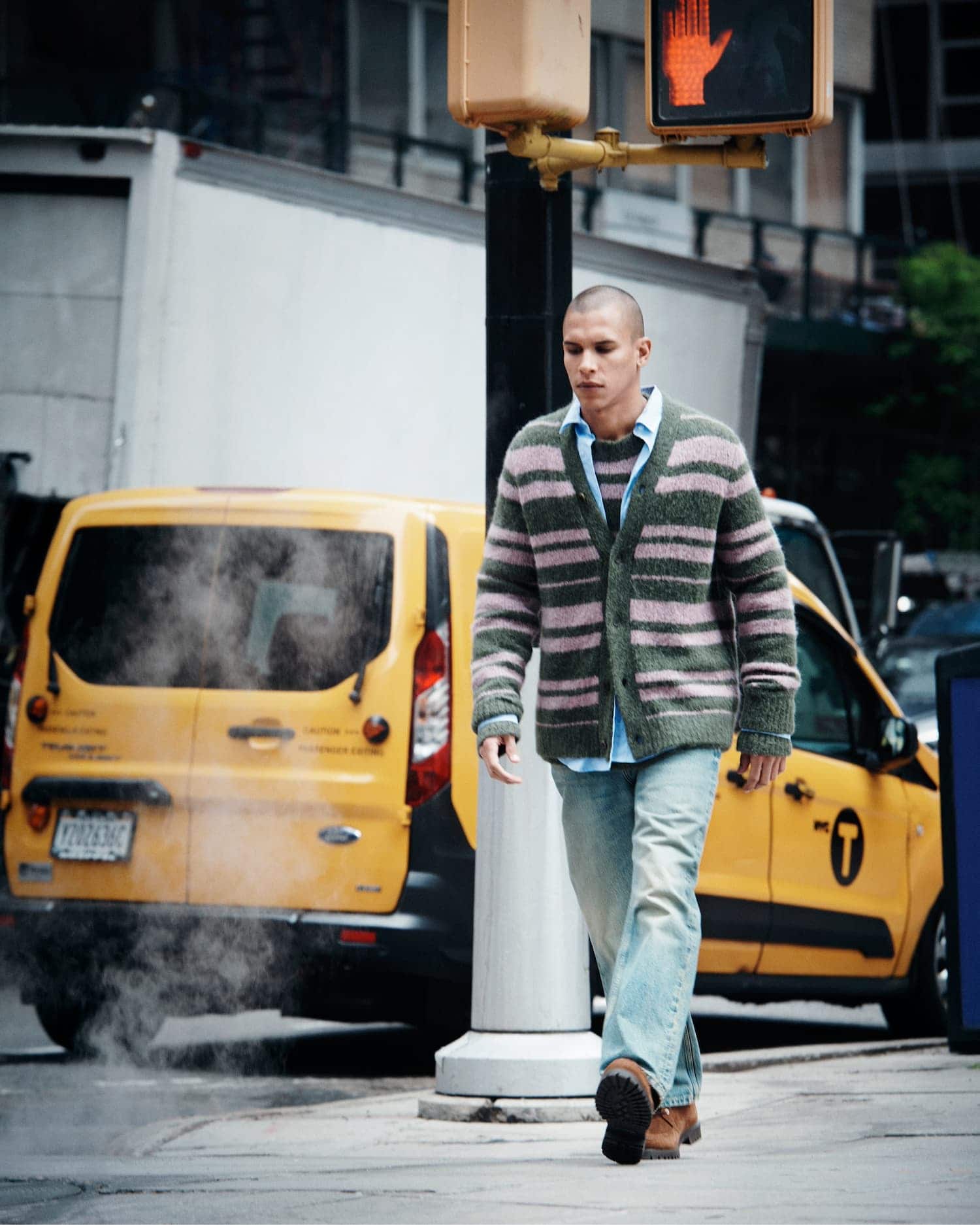 Man wearing a striped cardigan and light wash jeans walking down a city street past a yellow cab.