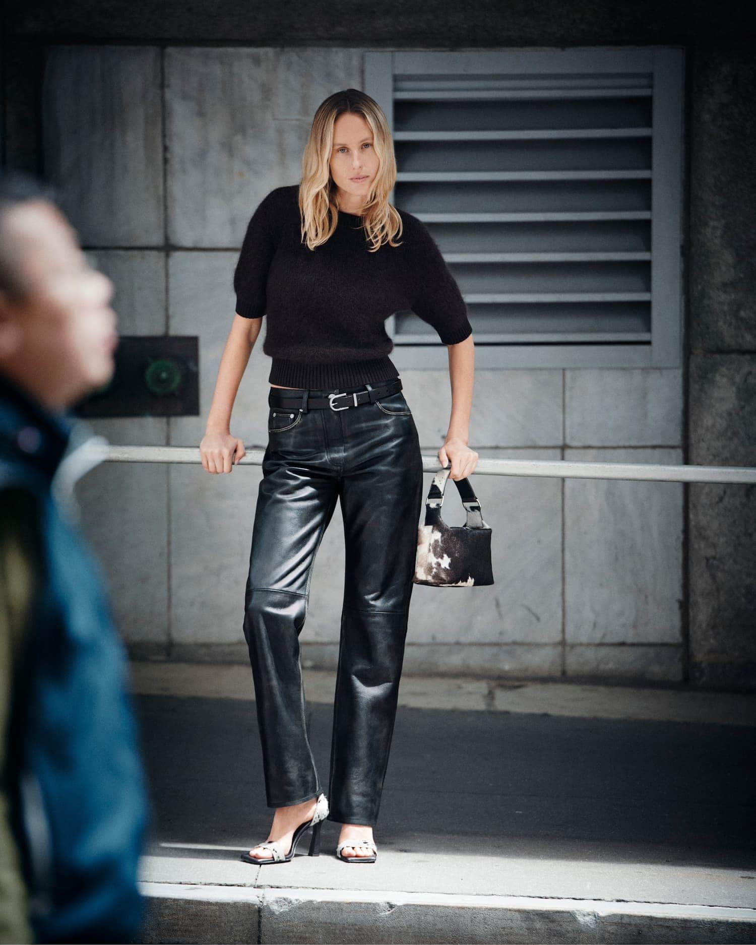 Model standing outdoors wearing a black sweater and leather trousers leaning against a metal railing