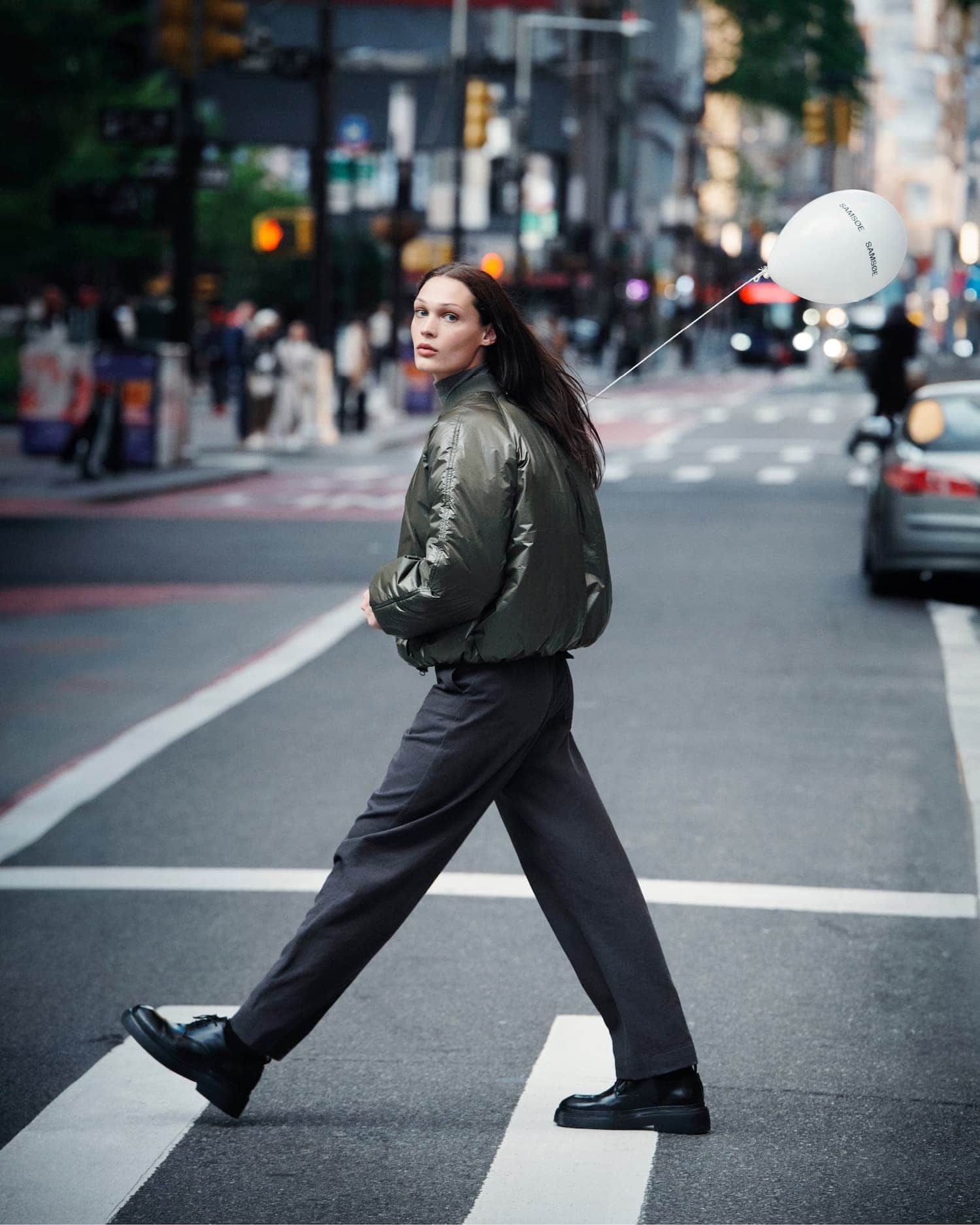 Model wearing a glossy olive bomber jacket walking across a New York City street holding a white balloon