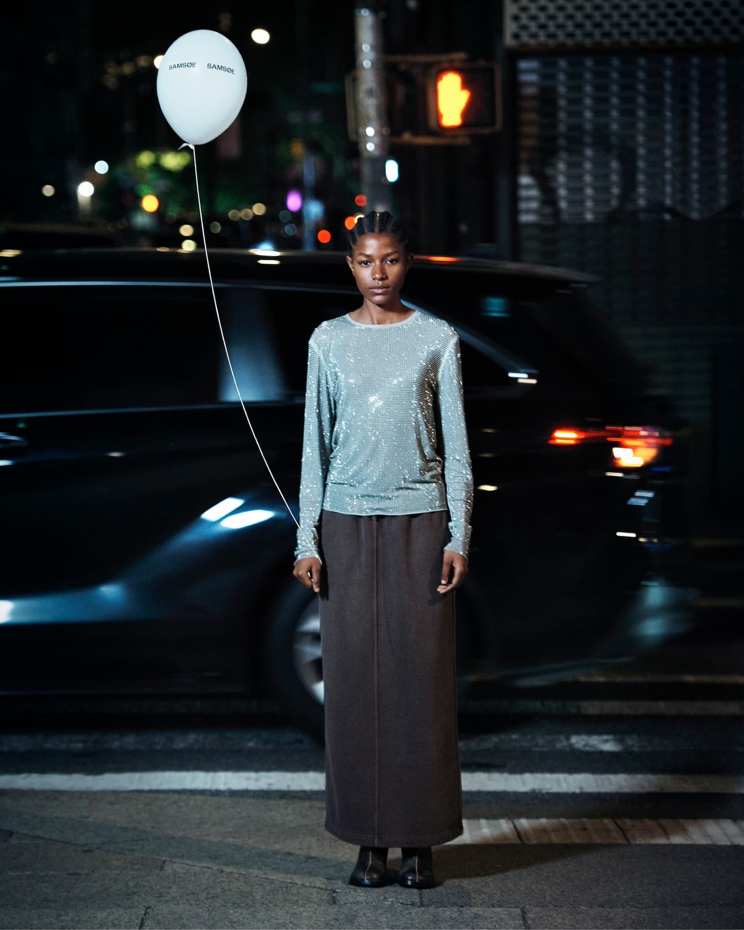 Model standing on a city street at night holding a white balloon next to a parked black car for Samsøe Samsøe