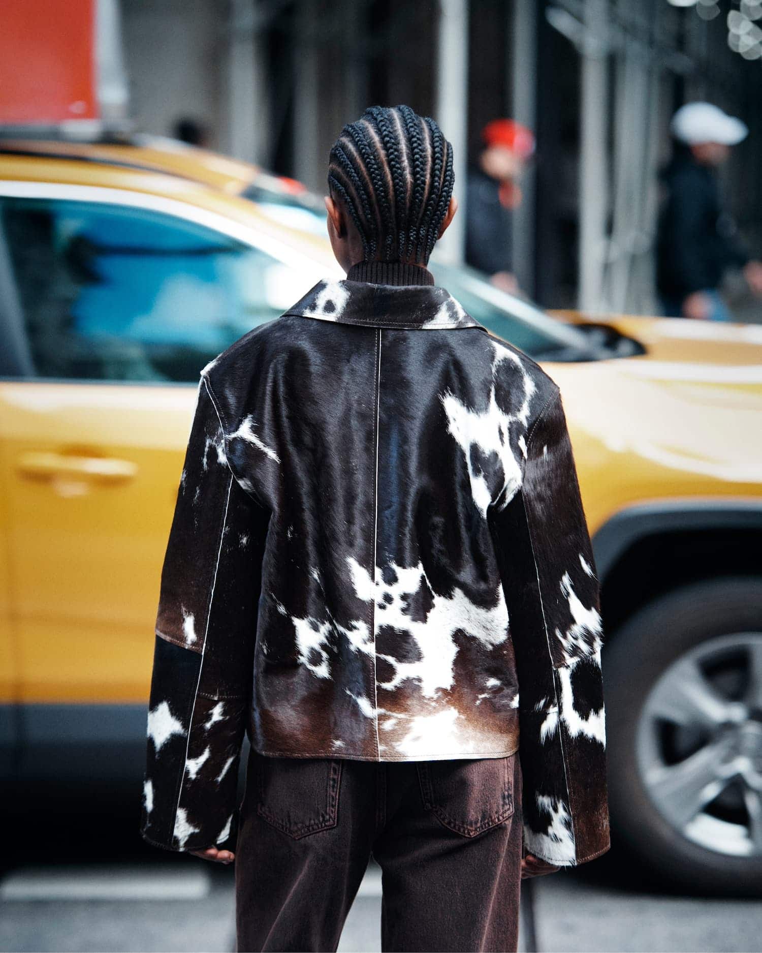 Model seen from behind wearing a cow-print leather jacket and dark trousers standing on a street in front of a yellow taxi.