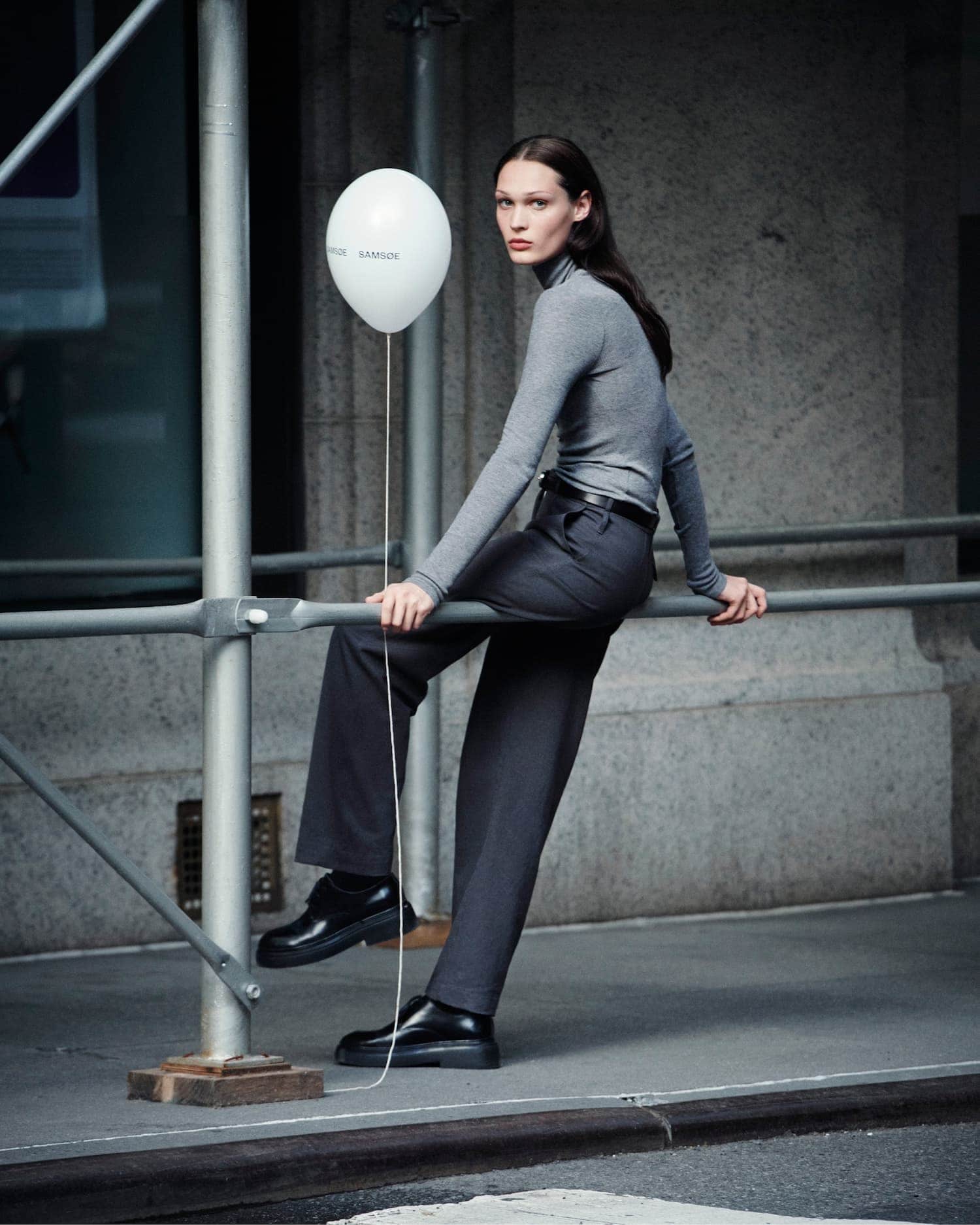 Model wearing a grey turtleneck and dark trousers holding a white balloon while leaning on scaffolding