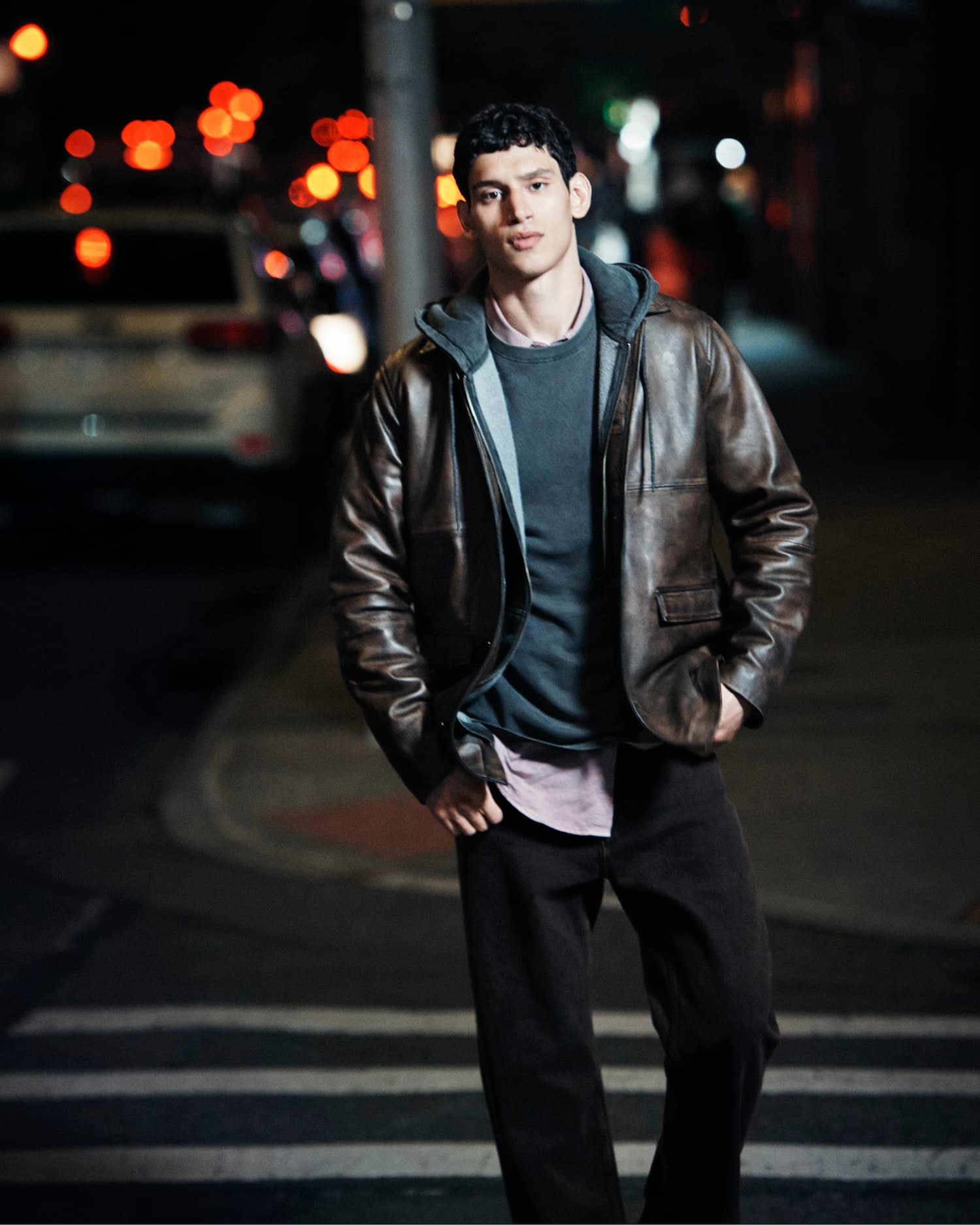 A man wearing a brown leather jacket and dark trousers standing on a city street at night with blurred traffic lights.
