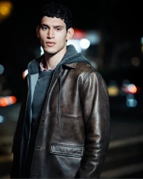 Man wearing a distressed brown leather jacket and grey hoodie against a dark nighttime street background