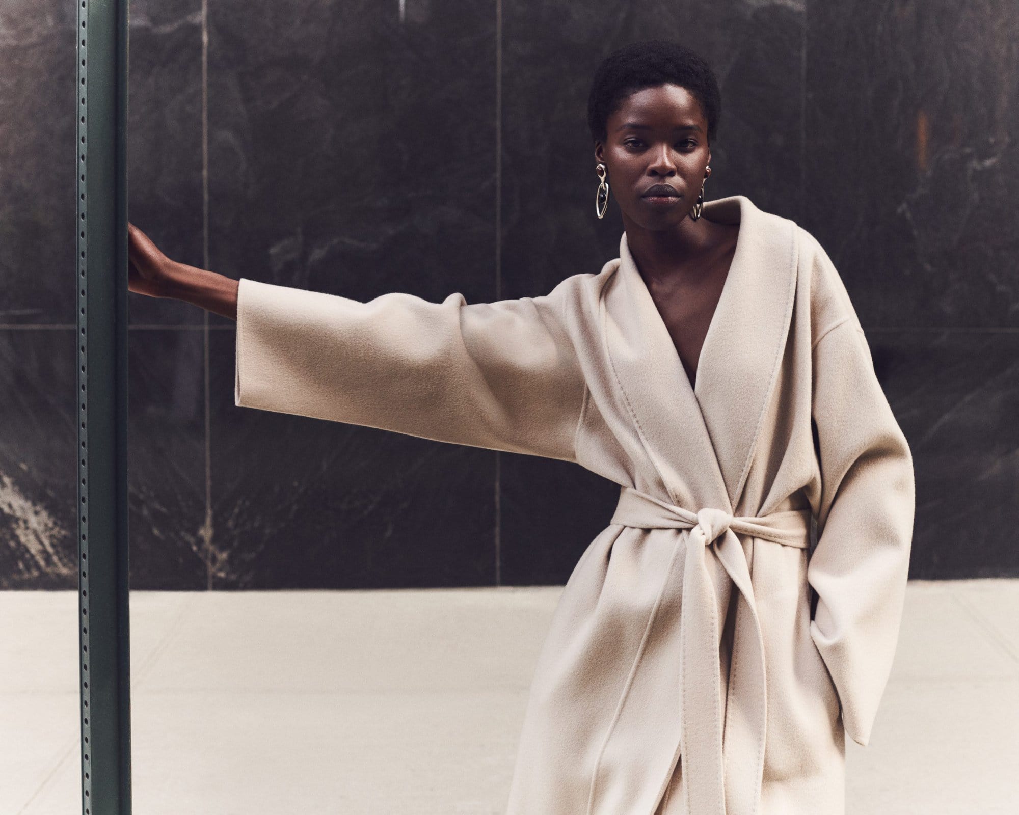 Model wearing a belted cream coat posing against a dark panel background for Lafayette 148