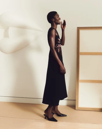 Black woman wearing a black sleeveless dress and heels laughing against a light studio wall with sculptural props.